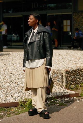 a photo of a woman wearing black platform sandals with pants layered with a skirt over with a button-down shirt and leather jacket