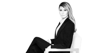 A high-contrast, black-and-white portrait of Melania Trump seated in a modern white chair against a stark white background, wearing a dark tailored suit and looking directly at the camera.
