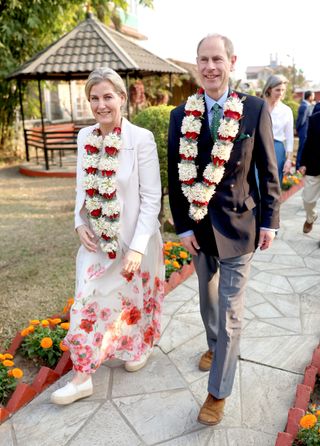 Duchess Sophie wearing a long white skirt with red flowers, a white blazer and a floral lei walking. next to Prince Edward wearing the same floral garland, a black blazer and gray pants walking along a sidewalk lined with orange flowers