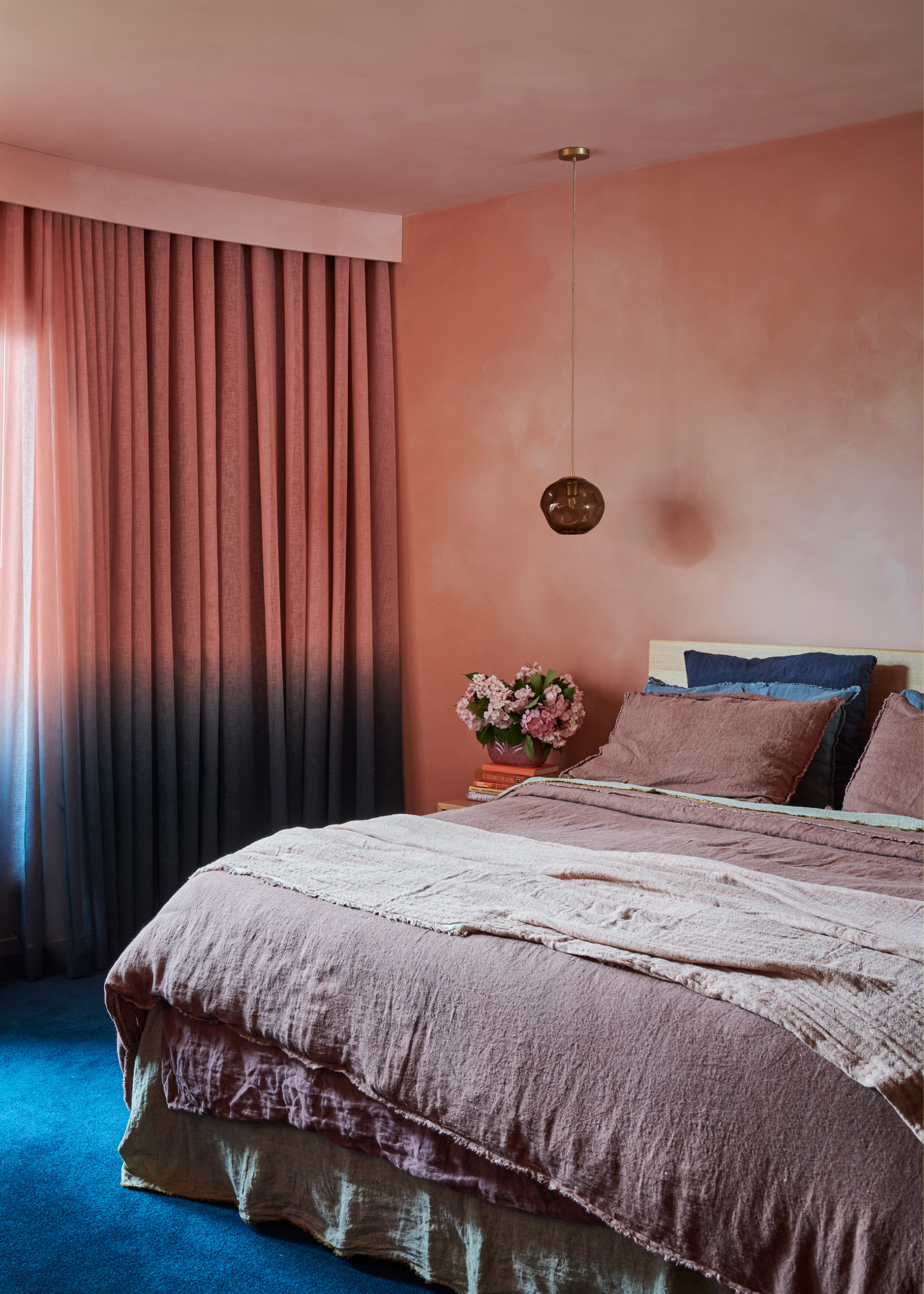A bedroom with blush walls, a pendant glass light, a planter with flowers, a layered bedscape with blush purple and blue pillows, and an ombre curtain