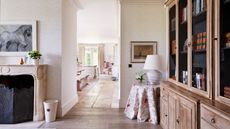 A bright living room with white walls, wooden cabinetry and a stone mantelpiece with a horse artwork above. Behind, through an open hallway, a kitchen is seen with bench seating and a kitchen island.