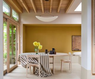 modern open dining space with dark yellow accent wall, wood ceiling beams, light flooring and light wood dining table and chairs