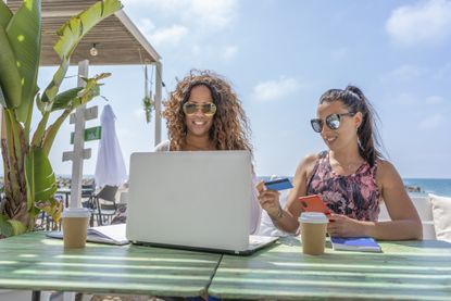 women friends shopping online through laptop using credit card whilst at a beach bar terrace
