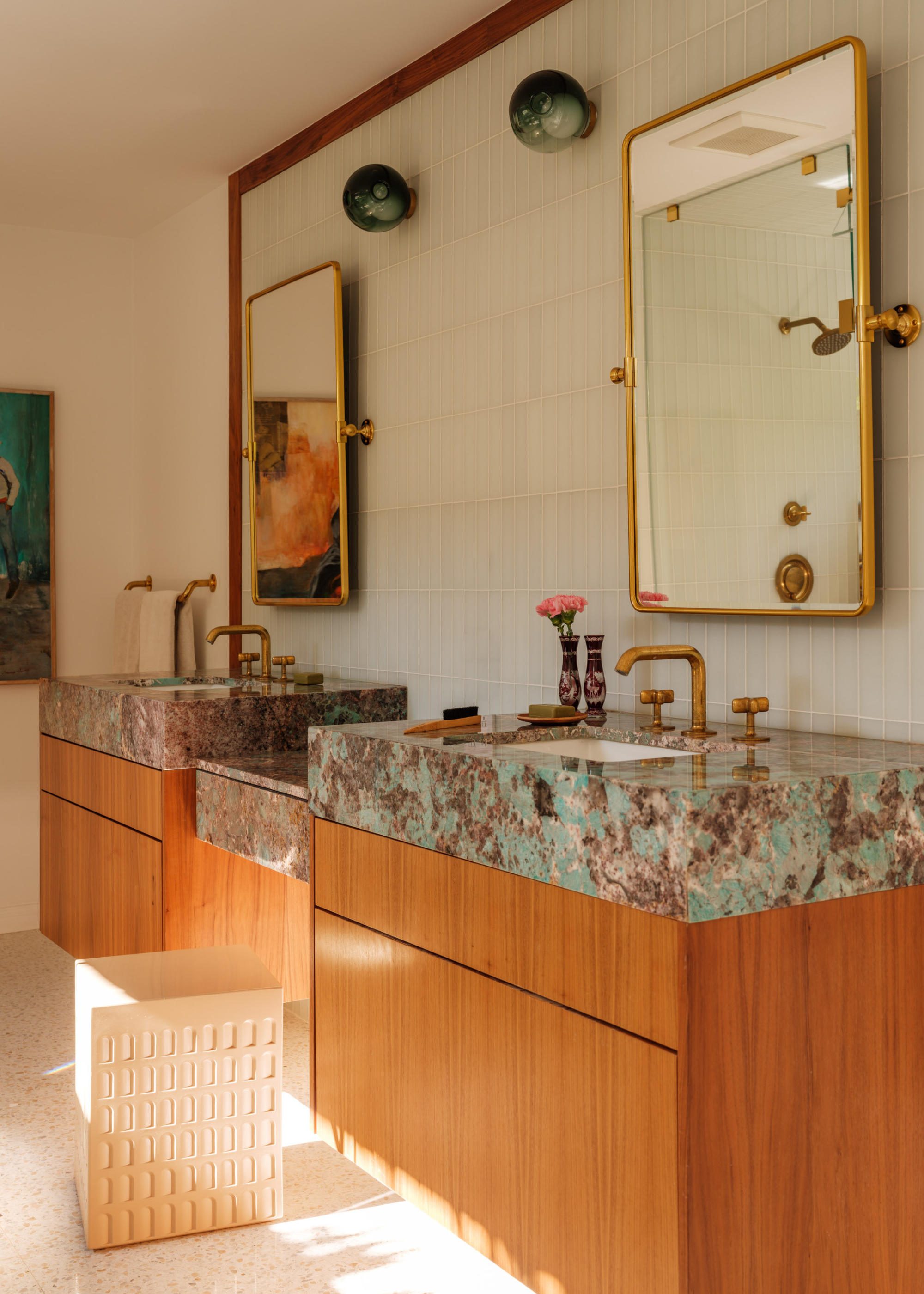 A his and hers bathroom with blue and gray stone countertops, wood storage drawers, a white seat/stool, gold mirrors, and brass hardware