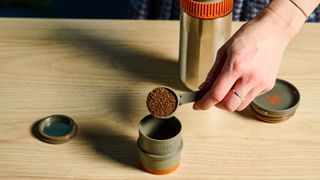 Scooping coffee into the Wacaco Pipamoka portable coffee maker in front of a blue background.