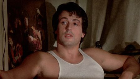 Rocky (Stallone) sits on the couch wearing a white tank top in Rocky (1977).