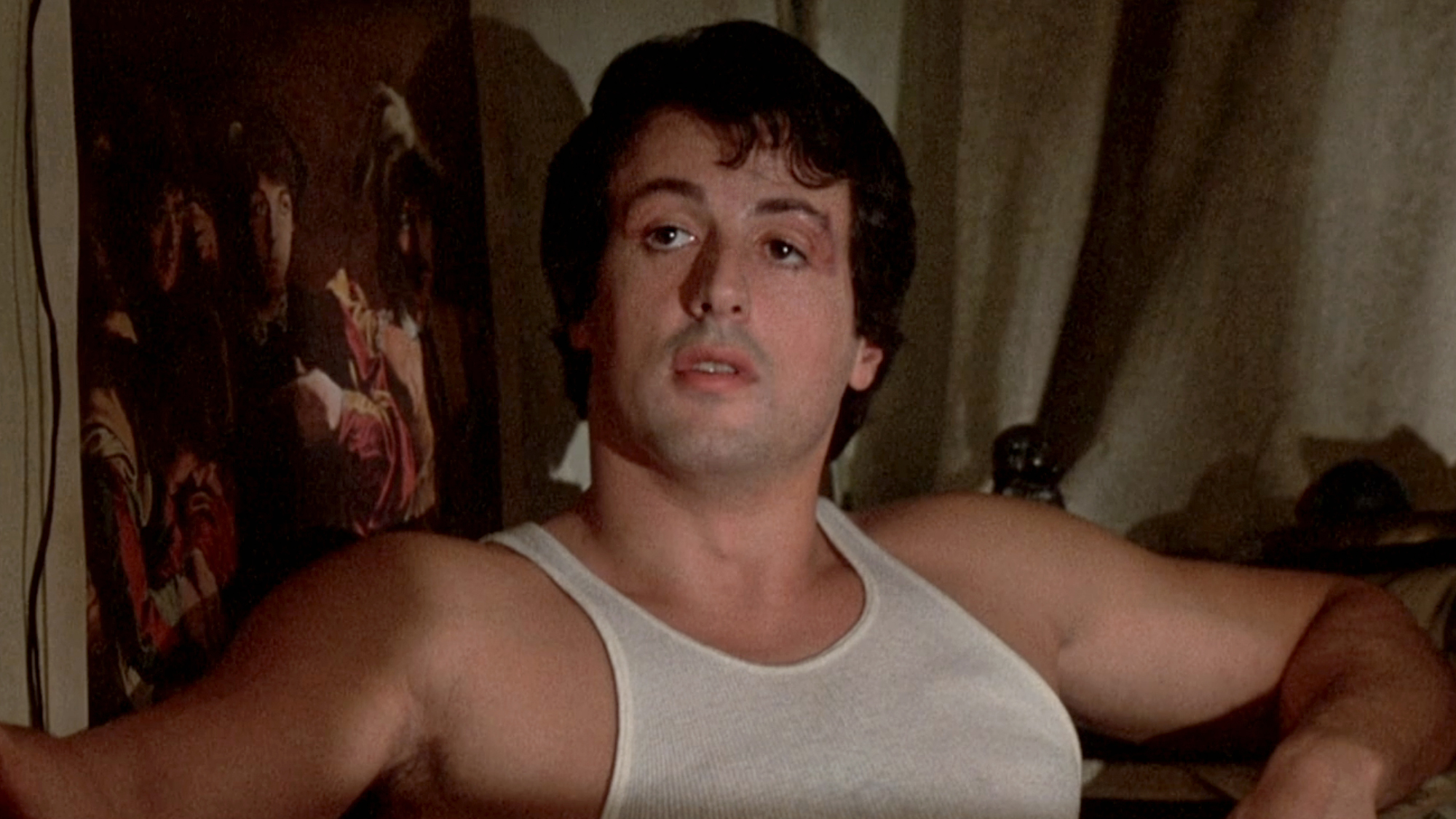 Rocky (Stallone) sits on the couch wearing a white tank top in Rocky (1977).