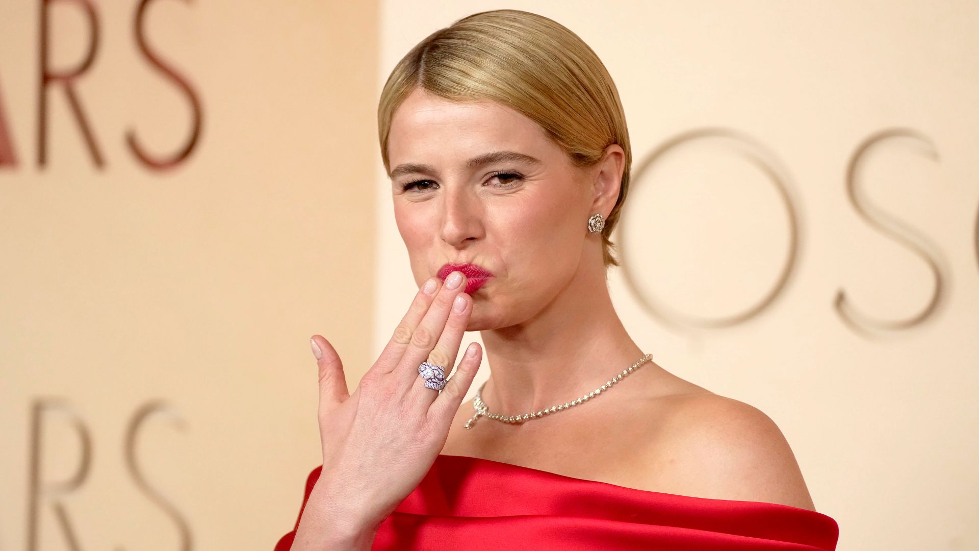 Jessie Buckley at the Oscars wearing a red dress with blonde cropped hair, blowing a kiss to the camera