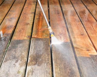 A pressure washer sprayer is cleaning a weathered treated wood deck. The background wood has been cleaned, the lower portion is dirty and weathered, showing the contrast