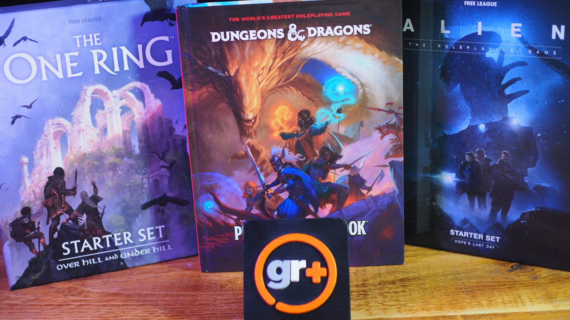 The One Ring Starter Set, Alien RPG Starter Set, and the D&amp;D Player's Handbook behind a GR+ logo on a wooden table