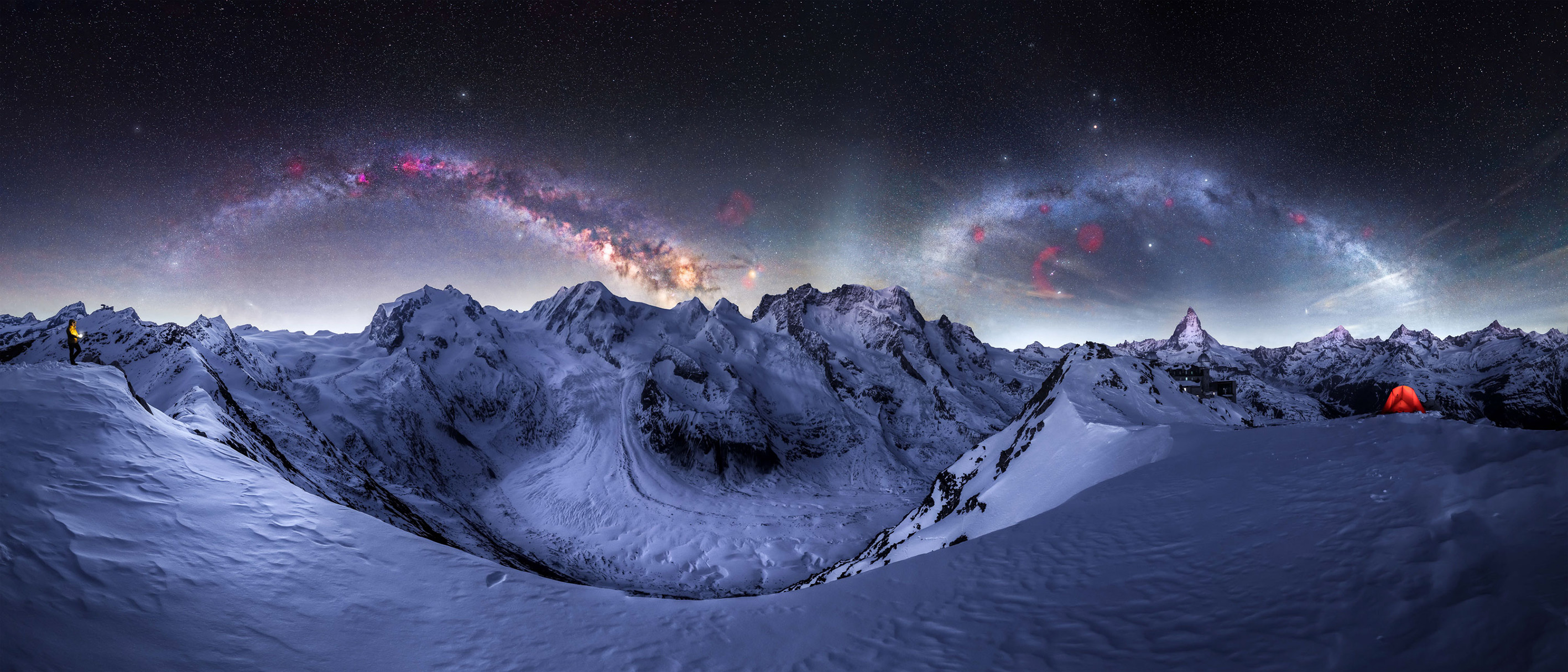 A panoramic view of snowy mountains under a starry sky, featuring the Milky Way and a lone figure beside a glowing red tent