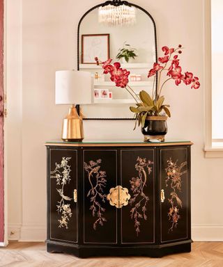 A white entryway with a black and gold console table with a gold lamp and flowers on it with a black mirror on the wall