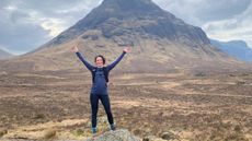 A woman trail runner on a rock holding her arms up with a Scottish mountain in the background