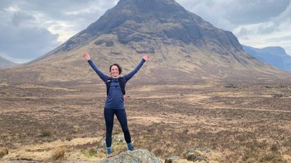 A woman trail runner on a rock holding her arms up with a Scottish mountain in the background