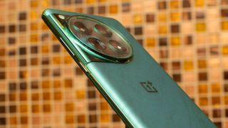 Baksidan och kameramodulen på en grön OnePlus 12 framför en gul/brun klinkervägg.
