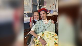 a young man seated and wearing a hospital gown and hat pictured next to his partner in a hospital room