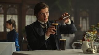 Louis Partridge pours a glass of Guinness out of a bottle while sat at a table