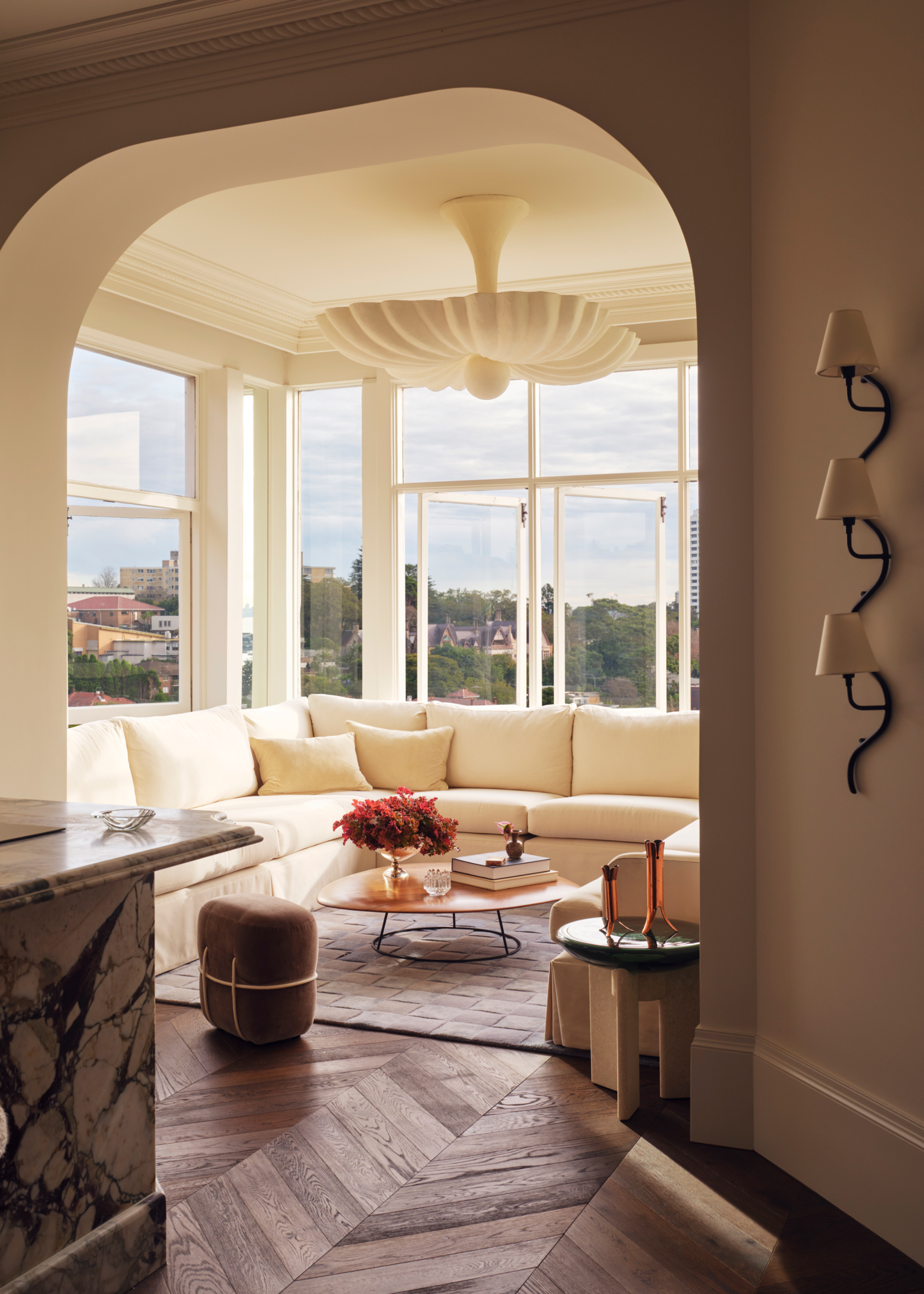 A bright conversation nook with a cream sofa, a low coffee table with books and a vase of stems, and a wall sconce with three lamps