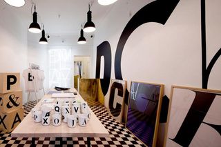 A black and white themed room. In the centre sits a long light wooden table on which sits many white mugs with black lettering. The floor is black and white chequered. The walls are white with black and white framed prints.
