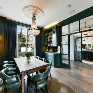 a dining room with dark green colour scheme with matching walls, curtains and display storage unit, with dark wooden floorboards and dining table with green leather chairs, opening out onto a hallway with a kitchen on the other side