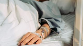 Close-up of a patients' hand while they are lying in a hospital bed. An IV drip is attached to their hand. 