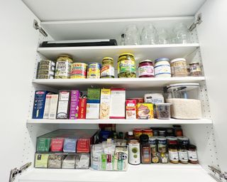 White organised food cupboard with clear containers with shelf-stable items such as herbs, pickled, and rice.