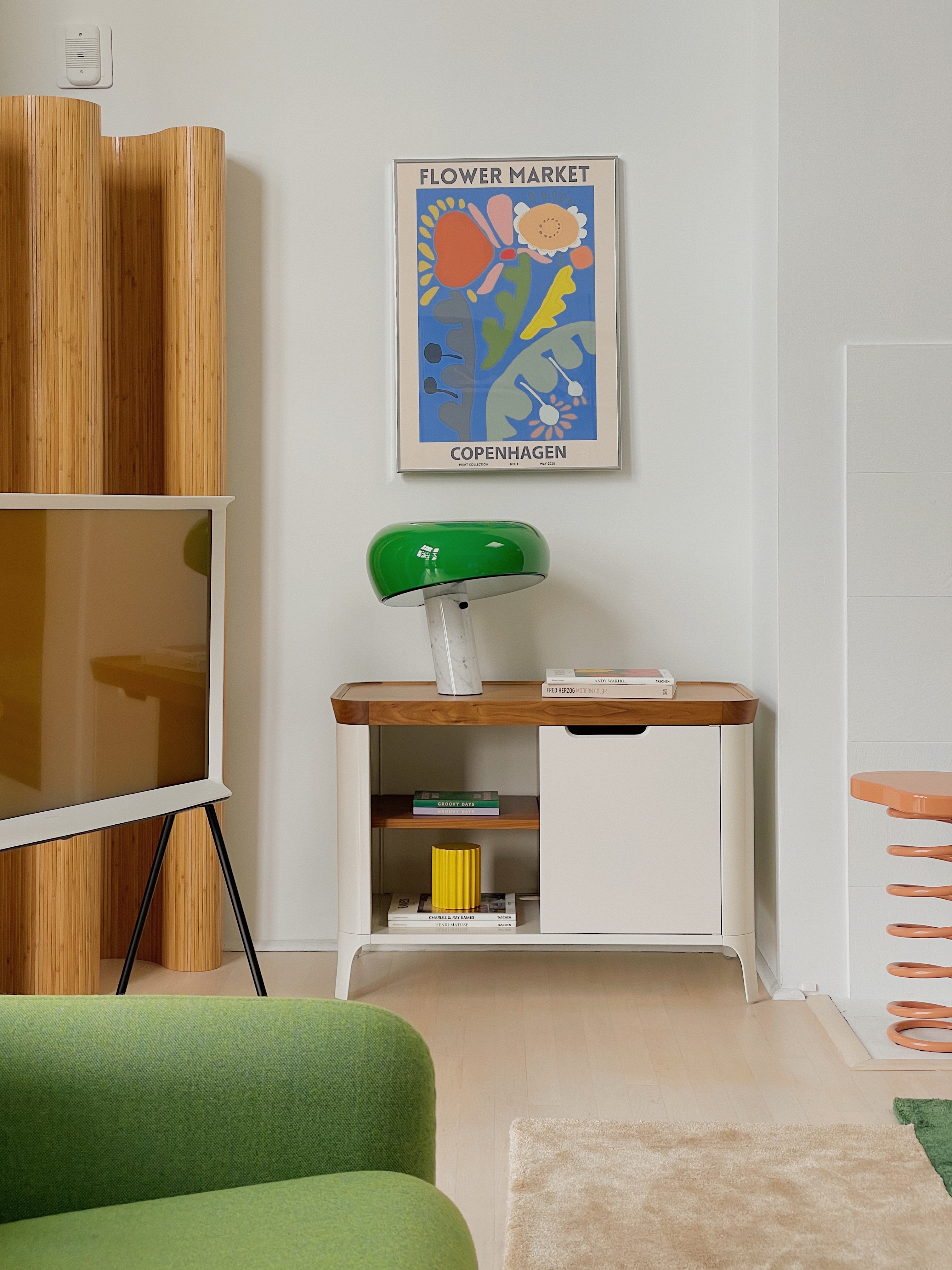 Image of a modern, white living room. There is the arm of a green sofa in view and a white cabinet with a wooden lid against the back wall. On the cabinet is a white and green mushroom lamp. To the left is a wooden, wavy room divider with a tv in front of it.