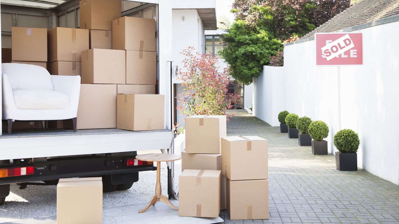 A large van with cardboard boxes and white accent chair unloaded outside house with pink sold sign