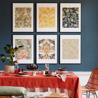 Dining table with red tablecloth, bench seat and chair on wooden floor, dark blue wall in the background with set if floral patterned pictures, framed wallpaper panels