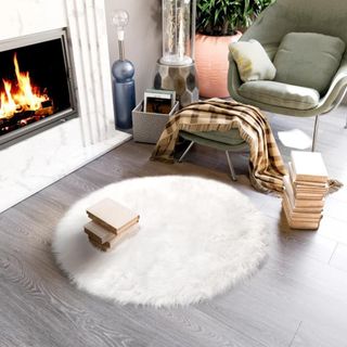 A round white faux fur rug sits next to a fireplace, a pile of books, and a gray suede chair