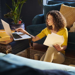 Woman wearing a yellow top, sat on the floor in front of a blue couch and a silver laptop. She is holding a piece of white paper and looking at the laptop screen