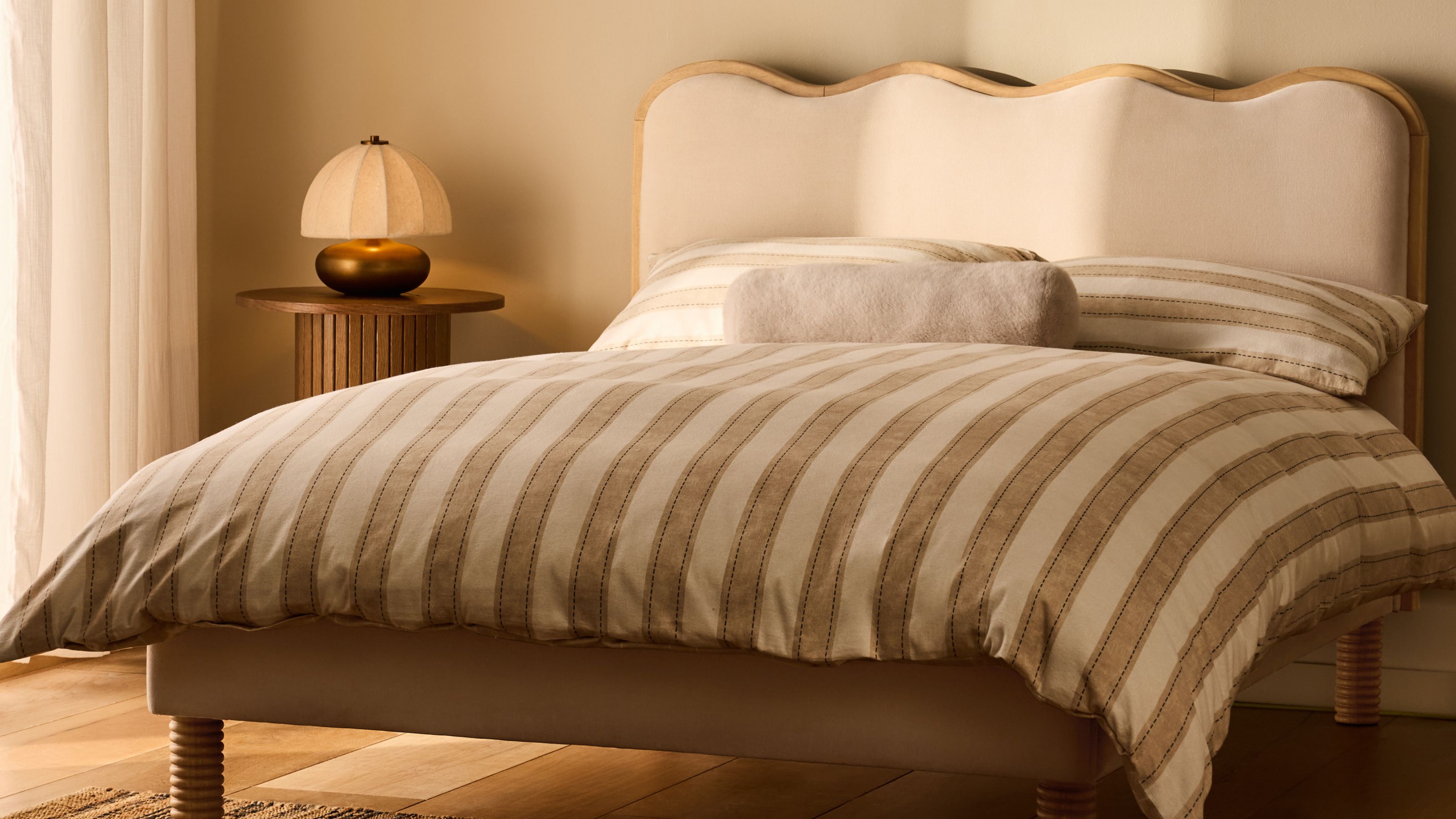 An off-white, wavy bedframe with a plain mattress on it in a beige bedroom. There is a wooden side table with a small table lamp on it.