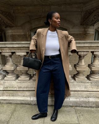 A woman wearing a white t-shirt, camel coat, black bag, blue barrel jeans, and black ankle boots.