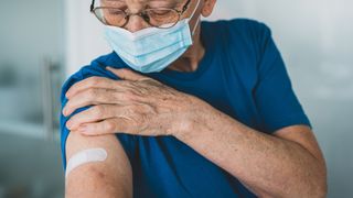 older man wearing a blue surgical mask looks down at his arm, which bears a bandage as if he's just gotten an injection