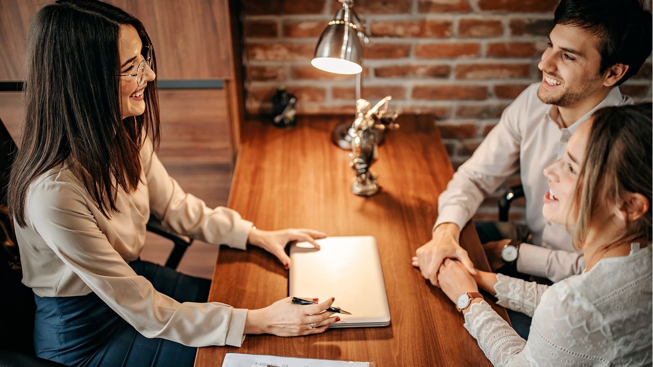 A young couple hold hands as they sit across from a smiling financial adviser.