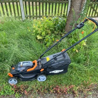 Worx WG779e cordless lawn mower on long grass next to tree and picket fence