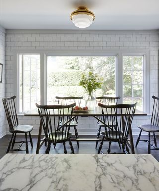 The dining area of a kitchen with white wall tiles, dark wood dining chair and large window