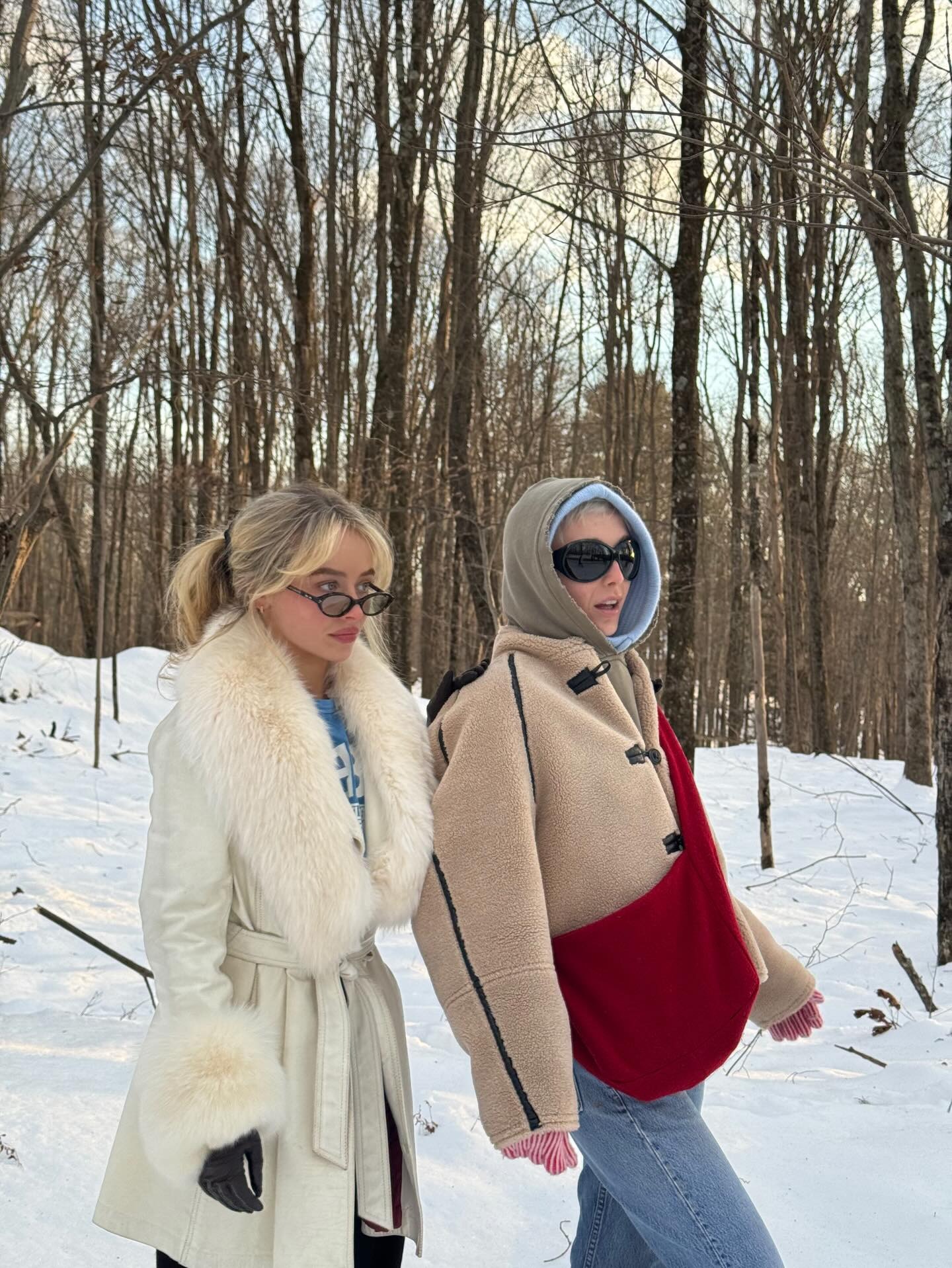 Sabrina Carpenter wears a white fur-trimmed coat over a blue t-shirt, standing next to Emma Chamberlina who wears a borge jacket with a khaki hood.