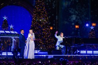 Alfonso Ribeiro and Julianne Hough stand to the left of Elton John as he waves from a piano. All three are on a Christmas tree-lined stage, in the THE WONDERFUL WORLD OF DISNEY: HOLIDAY SPECTACULAR.