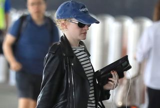 Julia Garner at LAX wearing a blue baseball cap, striped T-shirt, leather jacket, jeans, and Mary Jane flats.