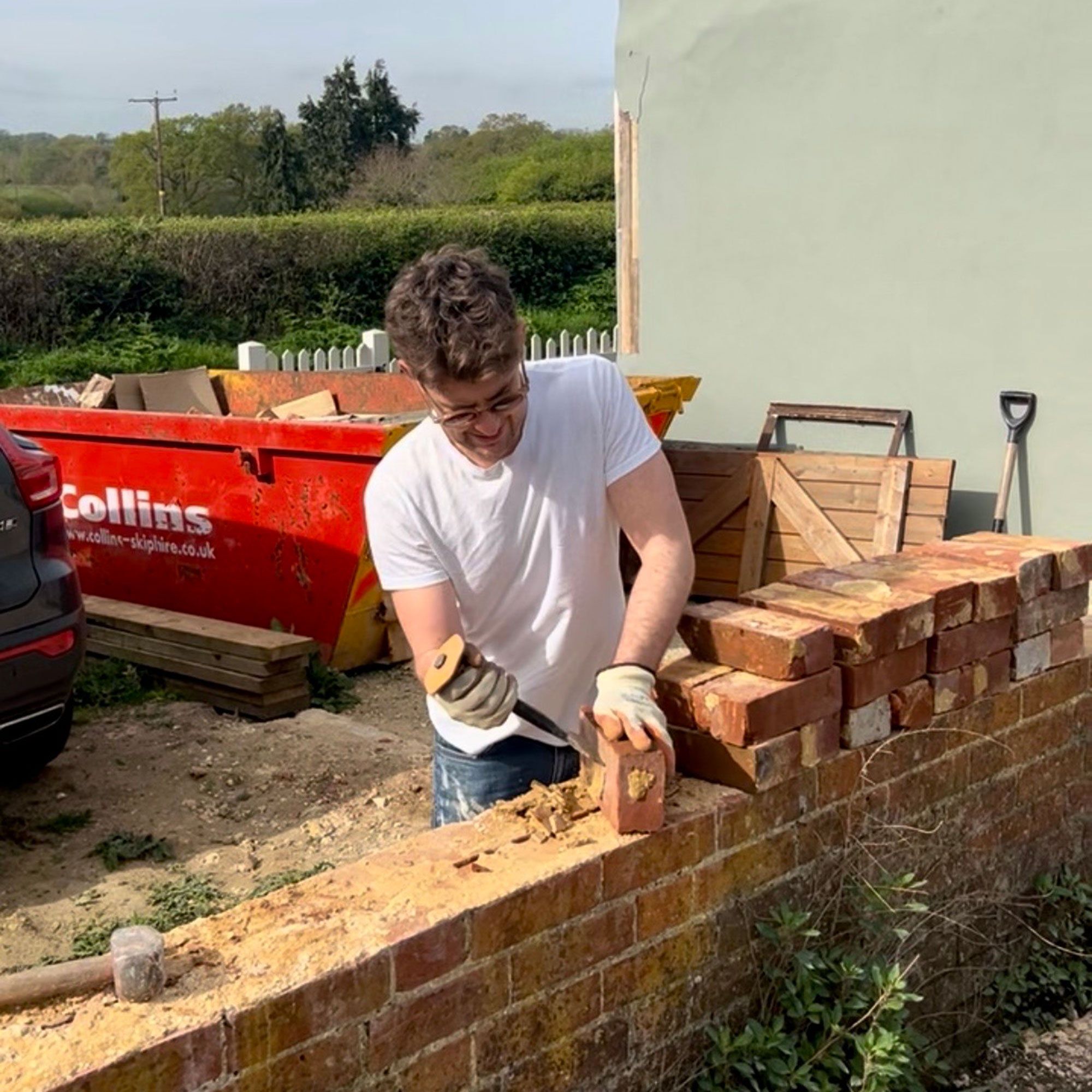 Man building outdoor brick wall with skip and country hedges in background