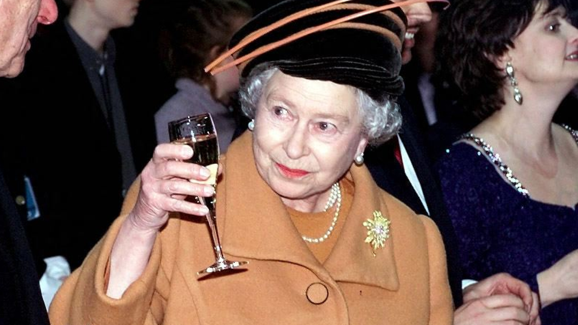 Queen Elizabeth wears an orange coat and raises a glass of champagne