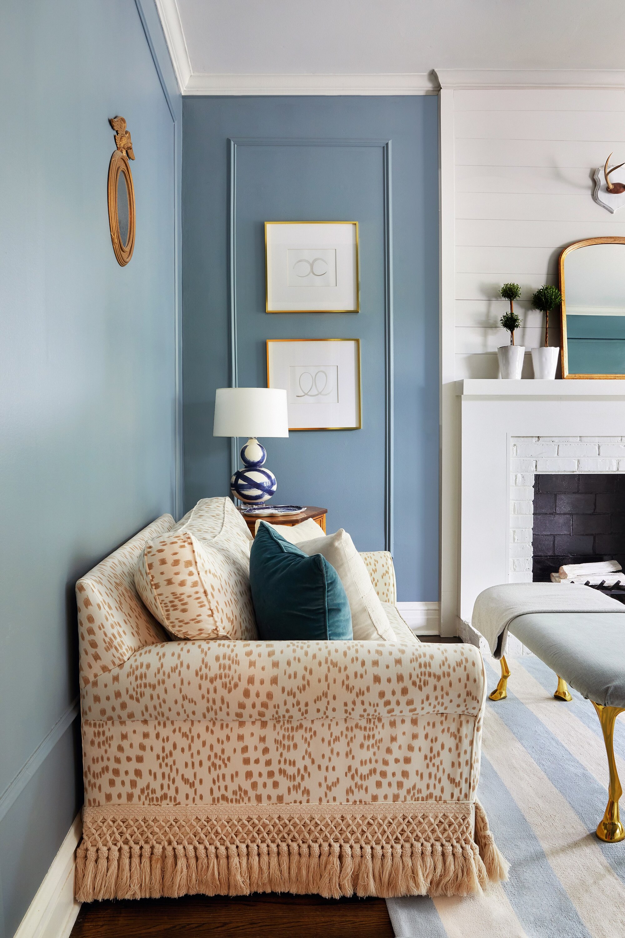 blue living room with white brick fireplace, ottoman, artwork on the walls with wall moldings, and a patterned sofa with a tasselled fringe