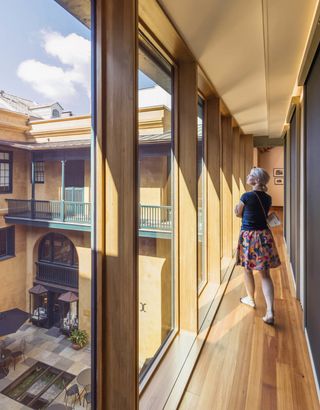 A woman dressed in a flowery skirt and blue short sleeve top walks along a wooden corridor filled with natural light in a yellow-hued building with green balconies.