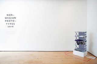 An open interior space, with a white wall featuring black text and a textile and metal shelf on a white box stand.
