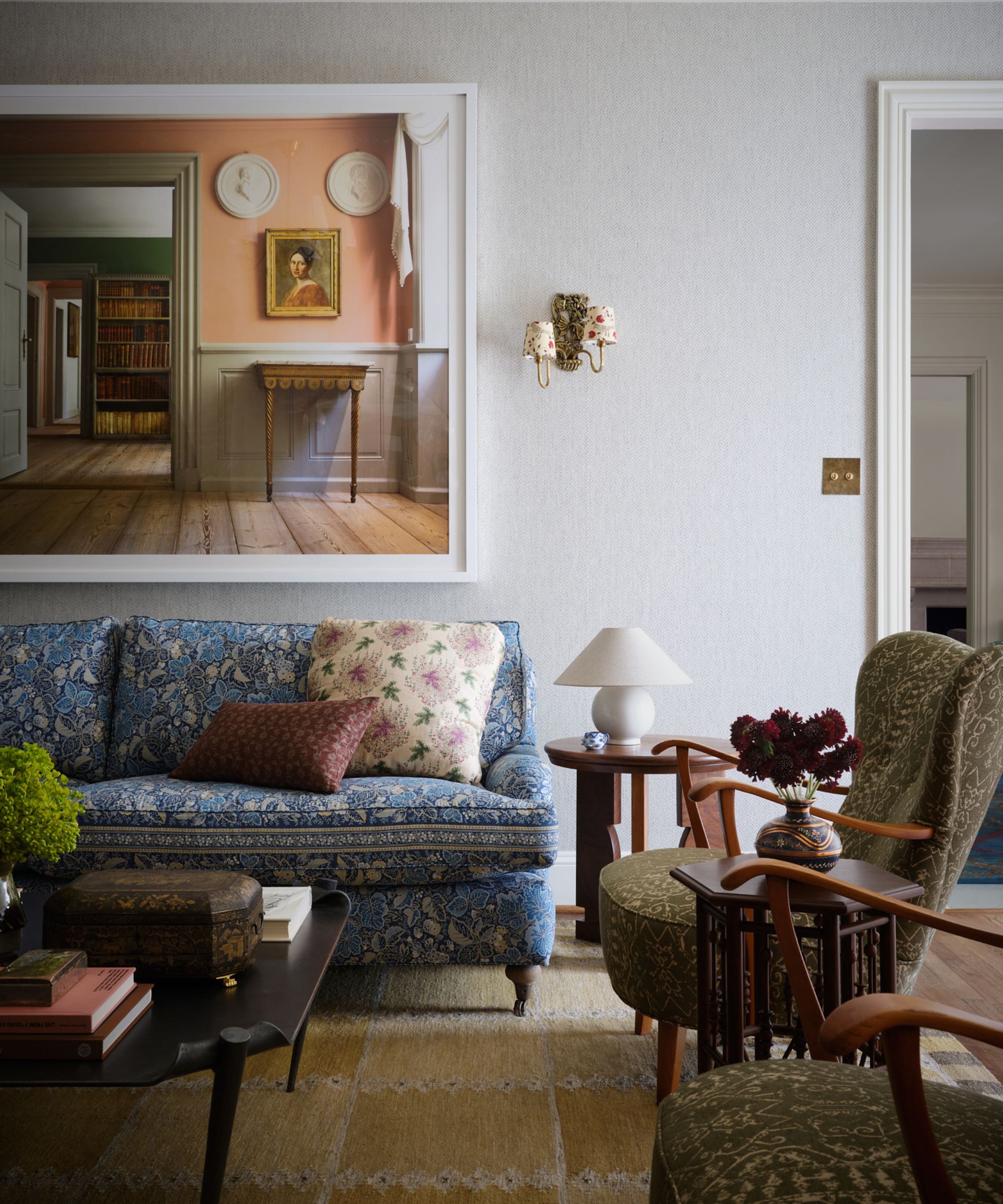 A living room with a blue patterned sofa, two vintage jacquard armchairs, a large print of a period property, a neutral rug, and a dark wood coffee table in the centre.