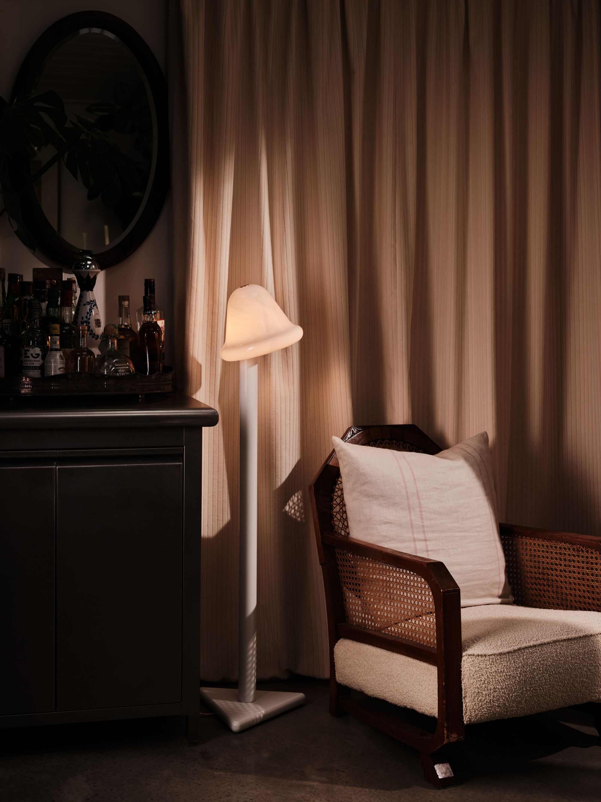 Image of a white floor lamp turned on in front of a curtain and beside a white boucle and dark wood accent chair.