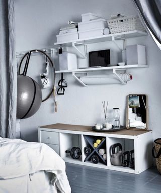 A home gym setup in a white bedroom with wall hooks, exercise ball and dumbbells stored in storage unit