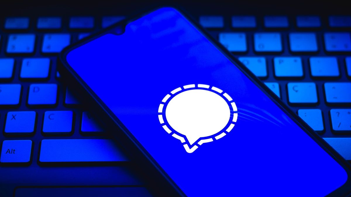 A smartphone sat on top of a white keyboard with the Signal app logo superimposed onto the phone&amp;#039;s display - an image all set in dark blue and black lighting