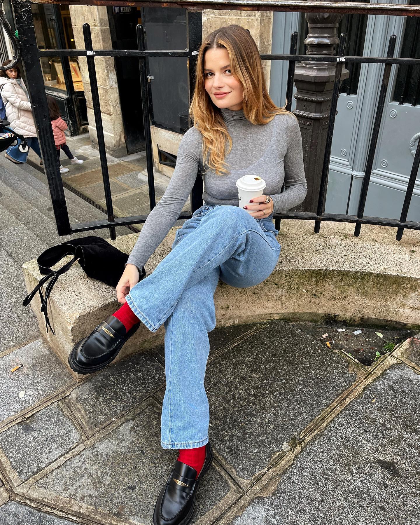 French influencer Sabina Socol sits on the streets of Paris wearing a gray turtleneck, jeans, red socks, and black loafers